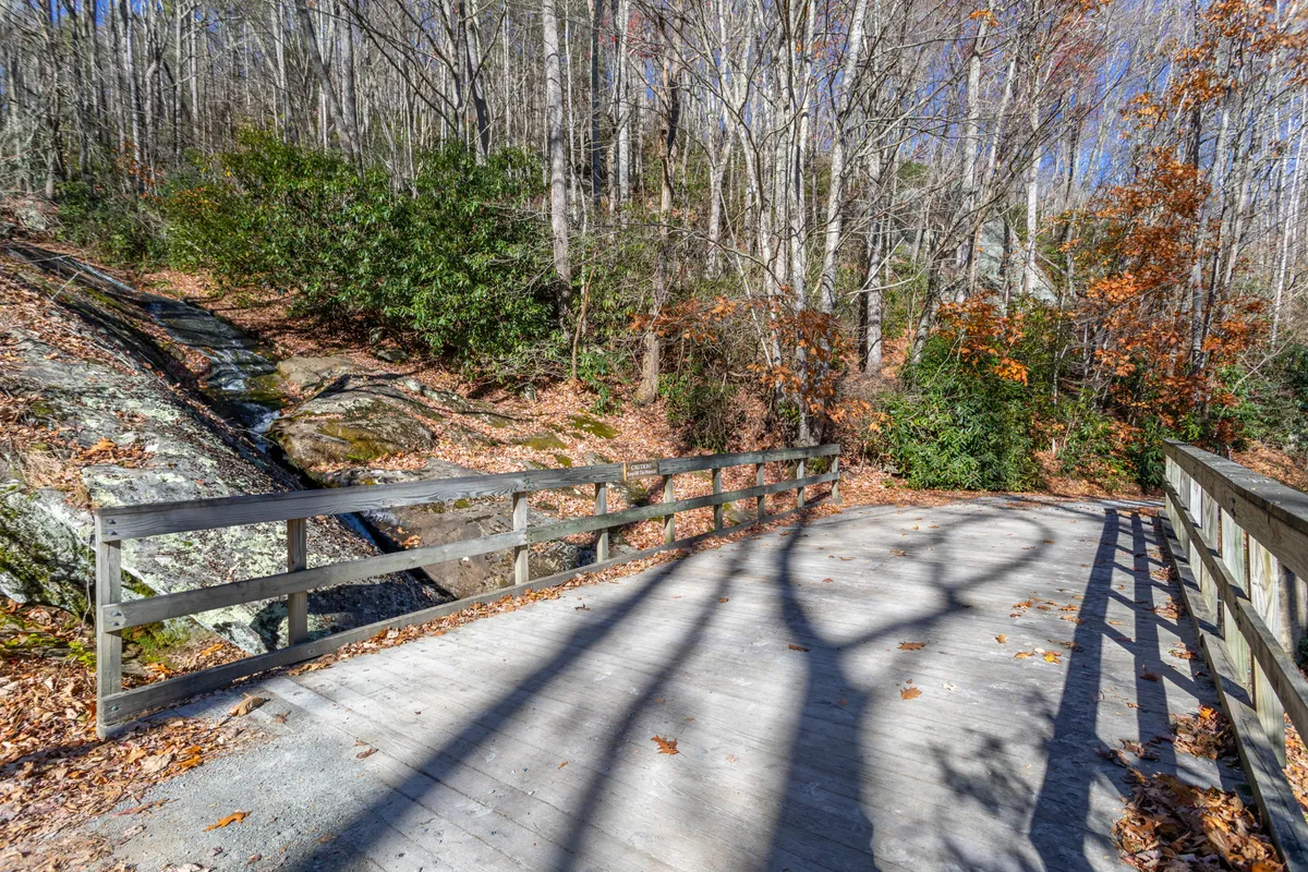 A charming wooden bridge crosses over a natural landscape, nestled among lush green trees and interesting rock formations. The setting feels secluded and storybook-like, inviting exploration and quiet moments by the water.
