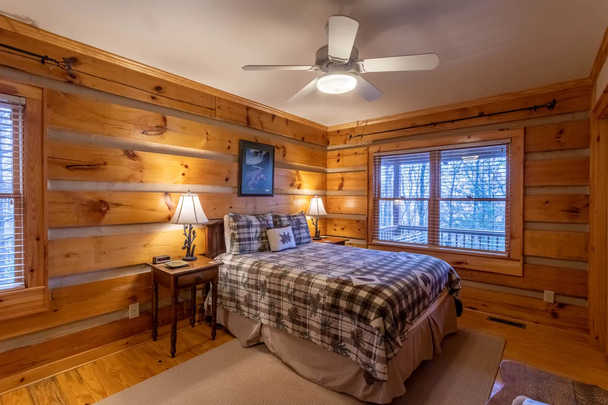 A tranquil bedroom with log walls, a plush bed dressed in plaid bedding, matching nightstands with soft-glowing lamps, and a large window inviting morning sunlight. The space exudes comfort and rustic charm, ideal for rest and rejuvenation after a day outdoors.