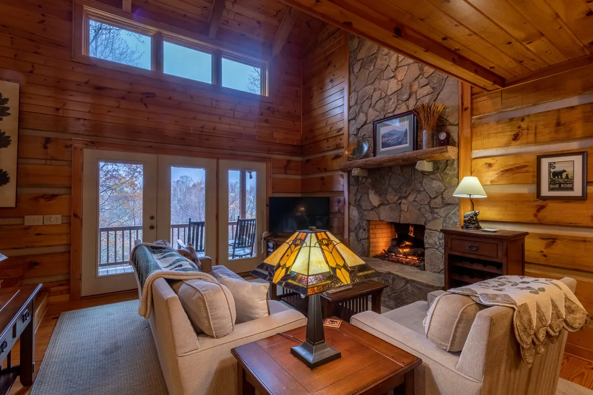 A cozy living space centered around a large stone fireplace, with plush sofas, stained wooden walls, a decorative lamp, and expansive windows offering glimpses of the outdoors. The ambiance is both rustic and comfortable, perfect for relaxation or socializing.