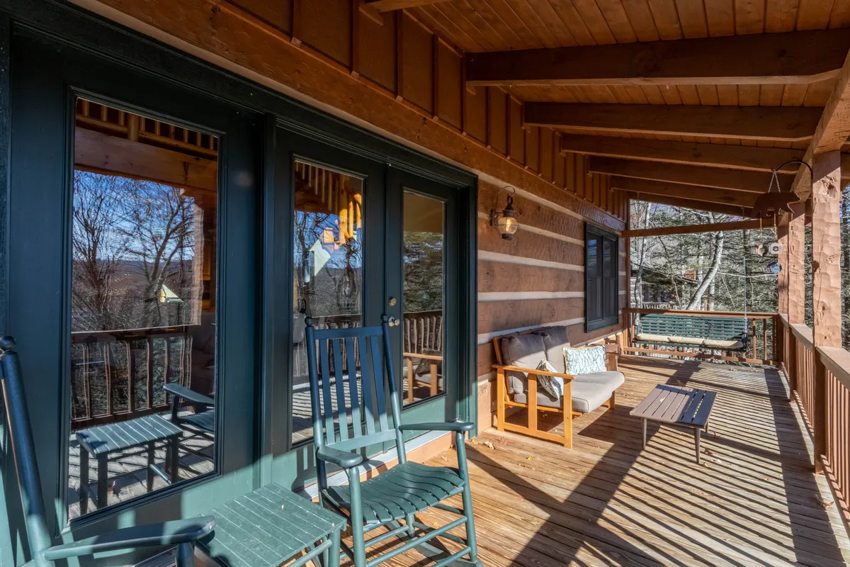 A cozy outdoor porch featuring rocking chairs, a wooden bench, and an outdoor table. Reflections in the windows and the view of the surrounding landscape invite couples to relax, enjoy sunsets, or listen to the sounds of nature together.