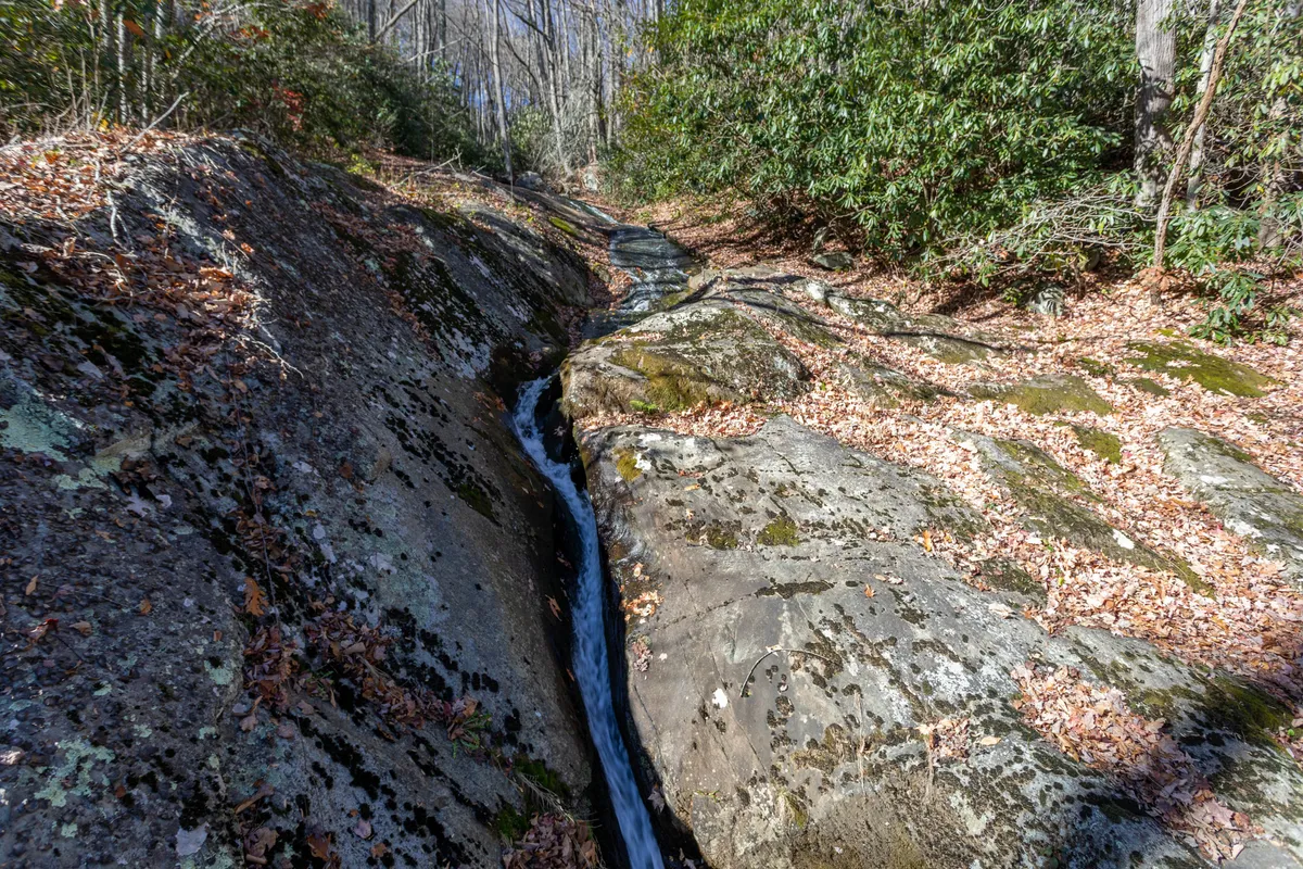 A rocky streambed with gently flowing water is surrounded by lush foliage, evoking the tranquil, rejuvenating atmosphere of a private mountain waterfall. The scene is peaceful, with the water's movement suggesting soothing sounds and a refreshing ambiance.