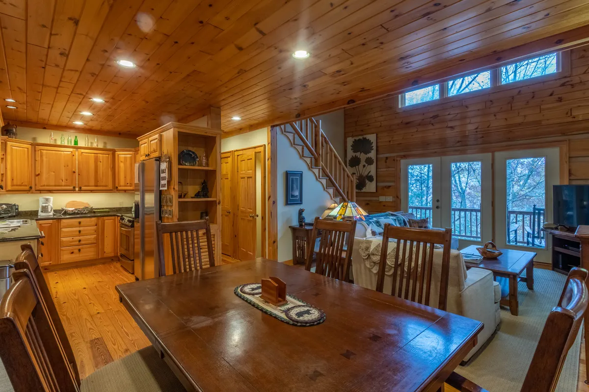 This cozy interior image showcases a wooden dining table, kitchen cabinetry, and large windows that suggest scenic views just outside. The space feels perfect for gathering and sharing meals, with an inviting ambiance and natural light pouring in.