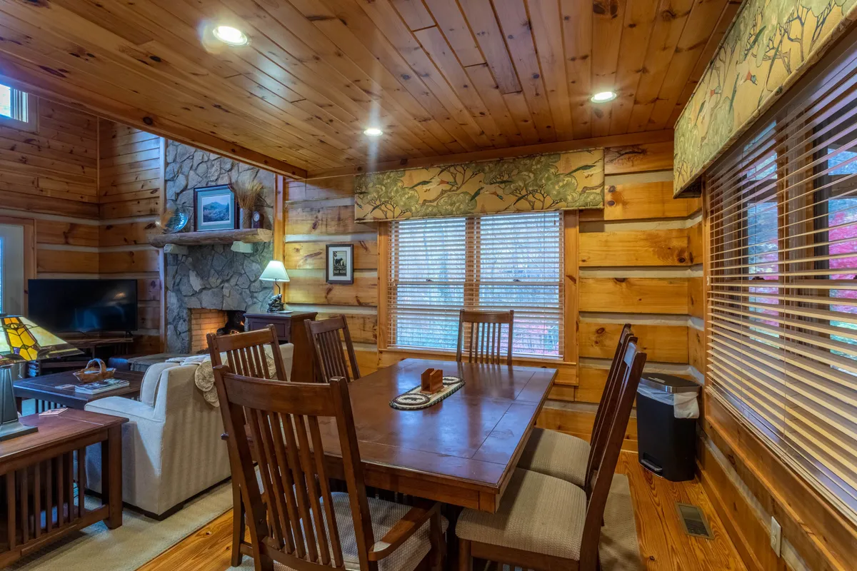 A dining area with a wooden table set near a fireplace, accented by wooden beams and window treatments. The setting is ideal for family meals, game nights, or holiday feasts, with a sense of warmth and togetherness.