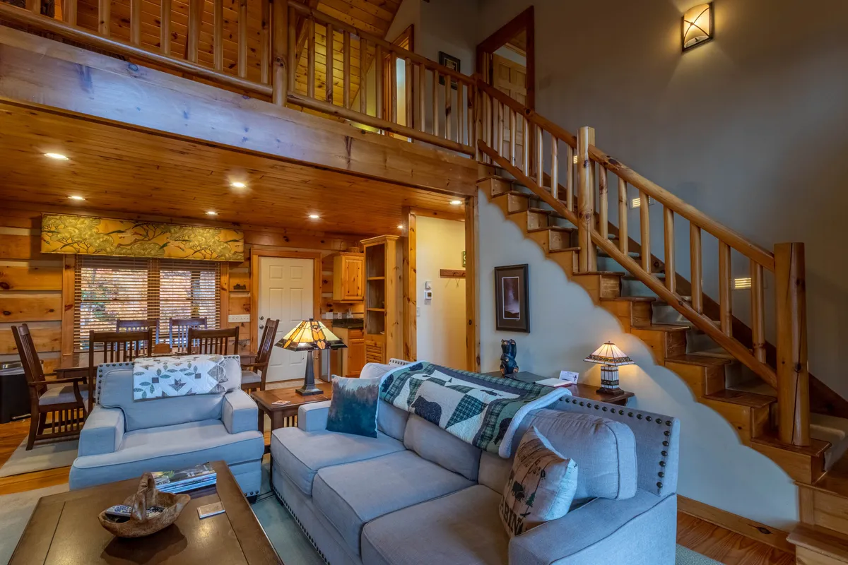A cozy living area featuring a plush sofa, a rustic wooden staircase, and a dining area warmly illuminated by soft lamps. The space invites groups to gather after a day outdoors, with comfortable seating and an atmosphere perfect for relaxation or storytelling by the fire.