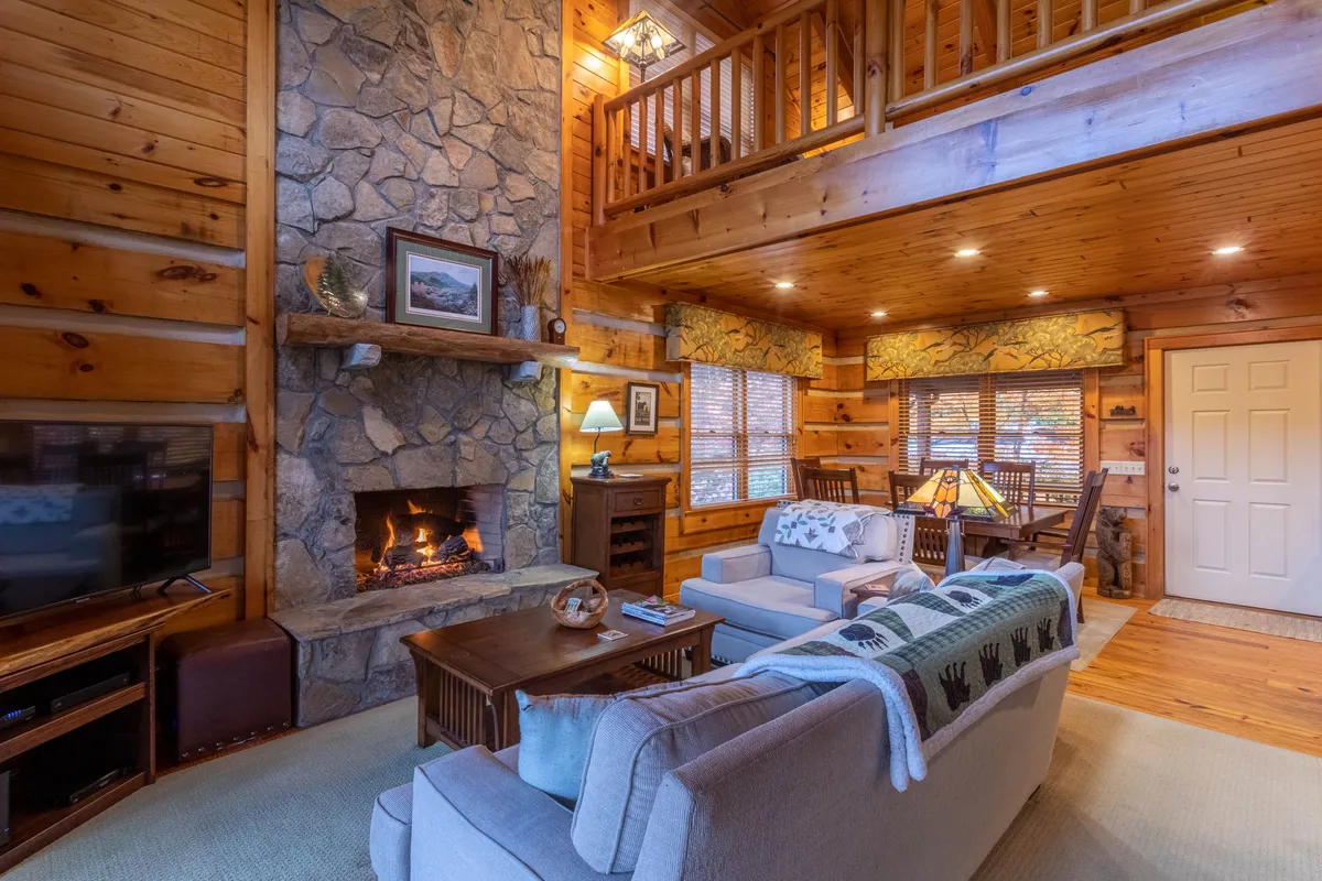 A spacious open-plan living and dining area featuring a prominent stone fireplace, comfortable seating, exposed wooden beams, and large windows that flood the space with natural light. The inviting arrangement encourages gathering, with the warmth of wood and stone accentuated by the sunlight streaming in.