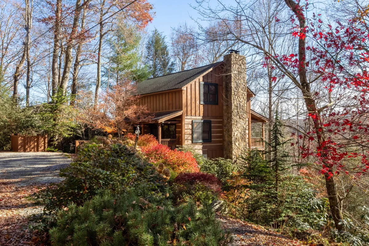 The image showcases a charming wooden cabin with a picturesque stone chimney, nestled among vibrant autumn foliage. The scene evokes a cozy, welcoming atmosphere perfect for relaxation, with hints of opportunities for outdoor gatherings or quiet evenings on the porch amid the brilliant colors of fall.