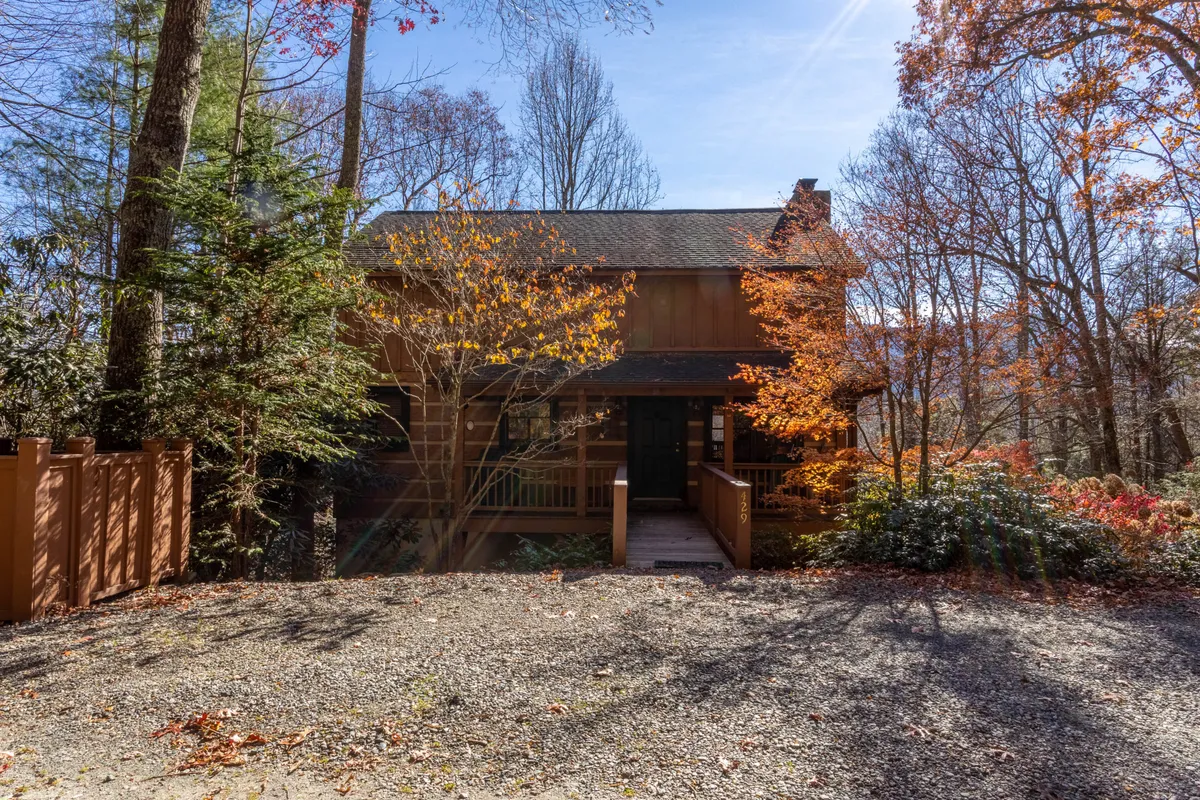 The rustic log cabin is embraced by vibrant autumn foliage, with a gravel driveway and a welcoming porch hinting at outdoor relaxation. The scene showcases the property’s connection to the surrounding landscape and the inviting atmosphere awaiting returning explorers.